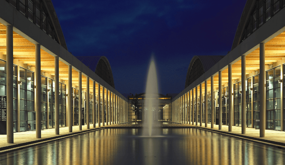 A nighttime view of a modern building featuring rows of tall columns and large glass windows, reflecting in a water pool. A tall fountain is visible in the center.
