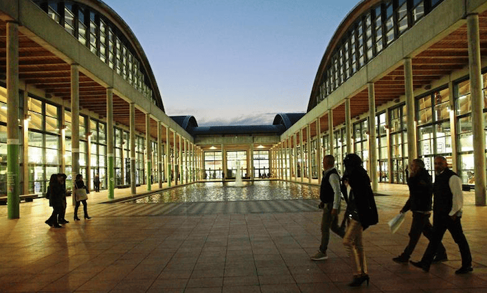 Wide view of a modern architectural space with glass walls and a water feature, featuring people walking during twilight.