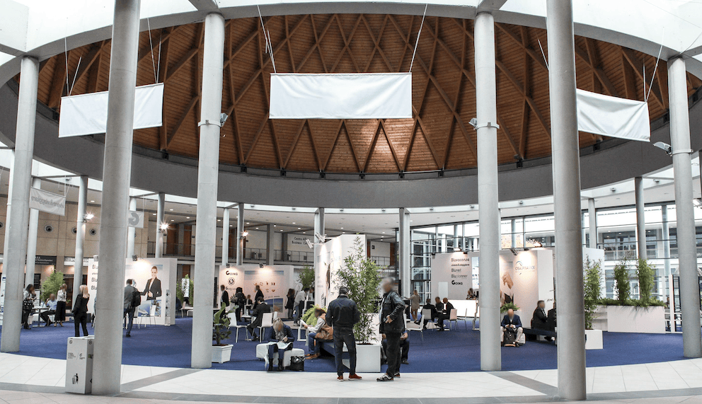 A spacious indoor event space featuring a circular design with a wooden ceiling. Various booths with displays and greenery are visible, along with people engaging in discussions and networking activities on a blue carpet.