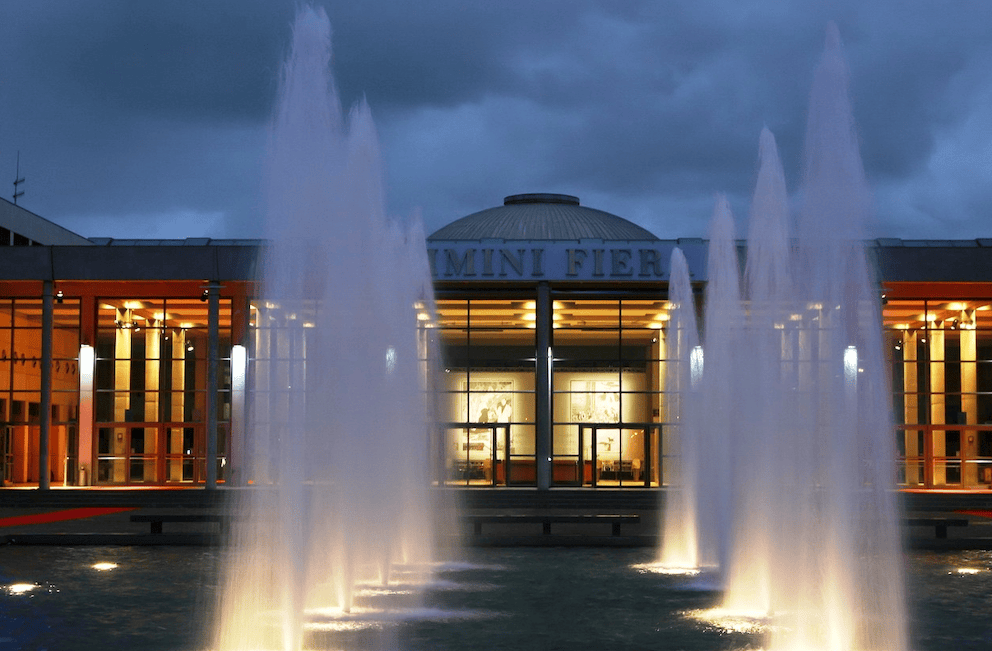 Illuminated fountains in front of a modern exhibition center at dusk, showcasing a welcoming entrance with bright lights and a glass facade.